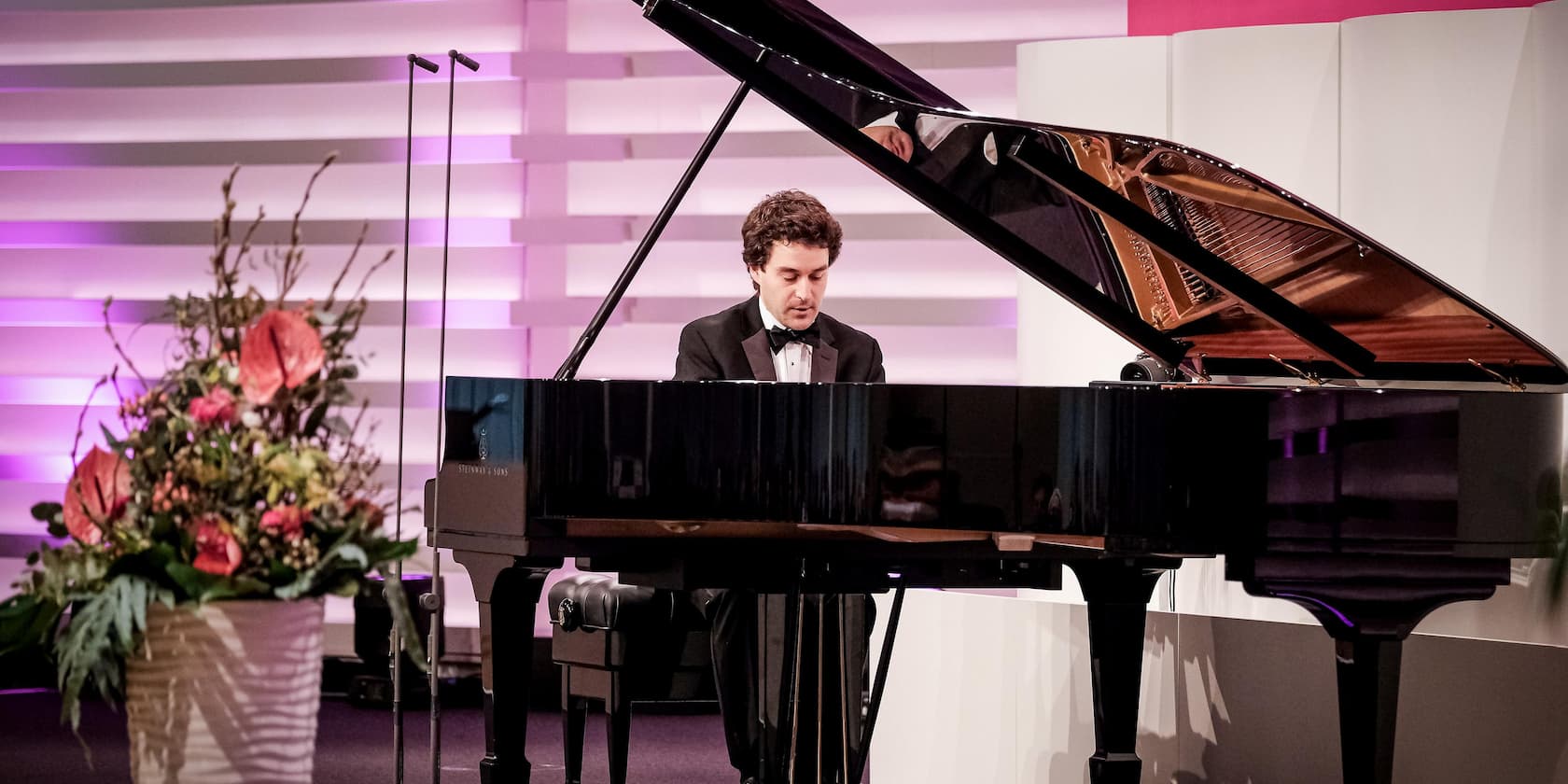 Ein Mann im Anzug spielt Klavier bei einem Konzert neben einem Blumenarrangement.