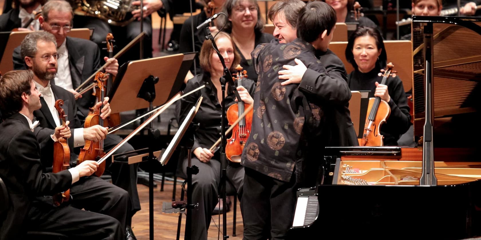 Ein Dirigent umarmt einen Musiker auf der Bühne, während das Orchester im Hintergrund steht.