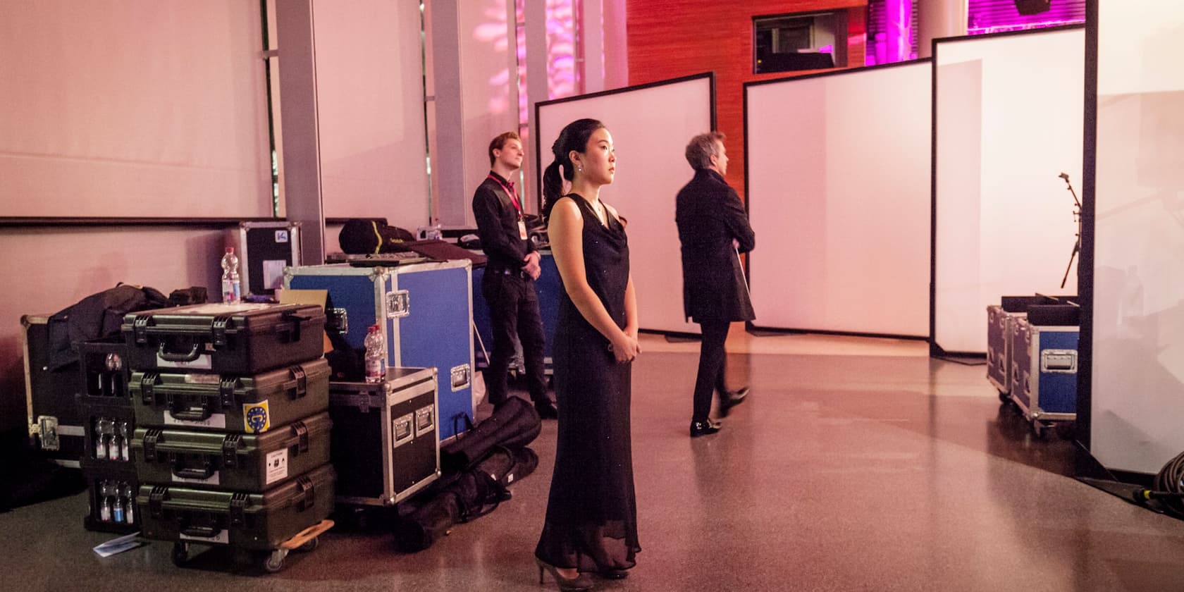 Woman in an elegant black dress stands backstage at a venue, surrounded by technical equipment and staff members.