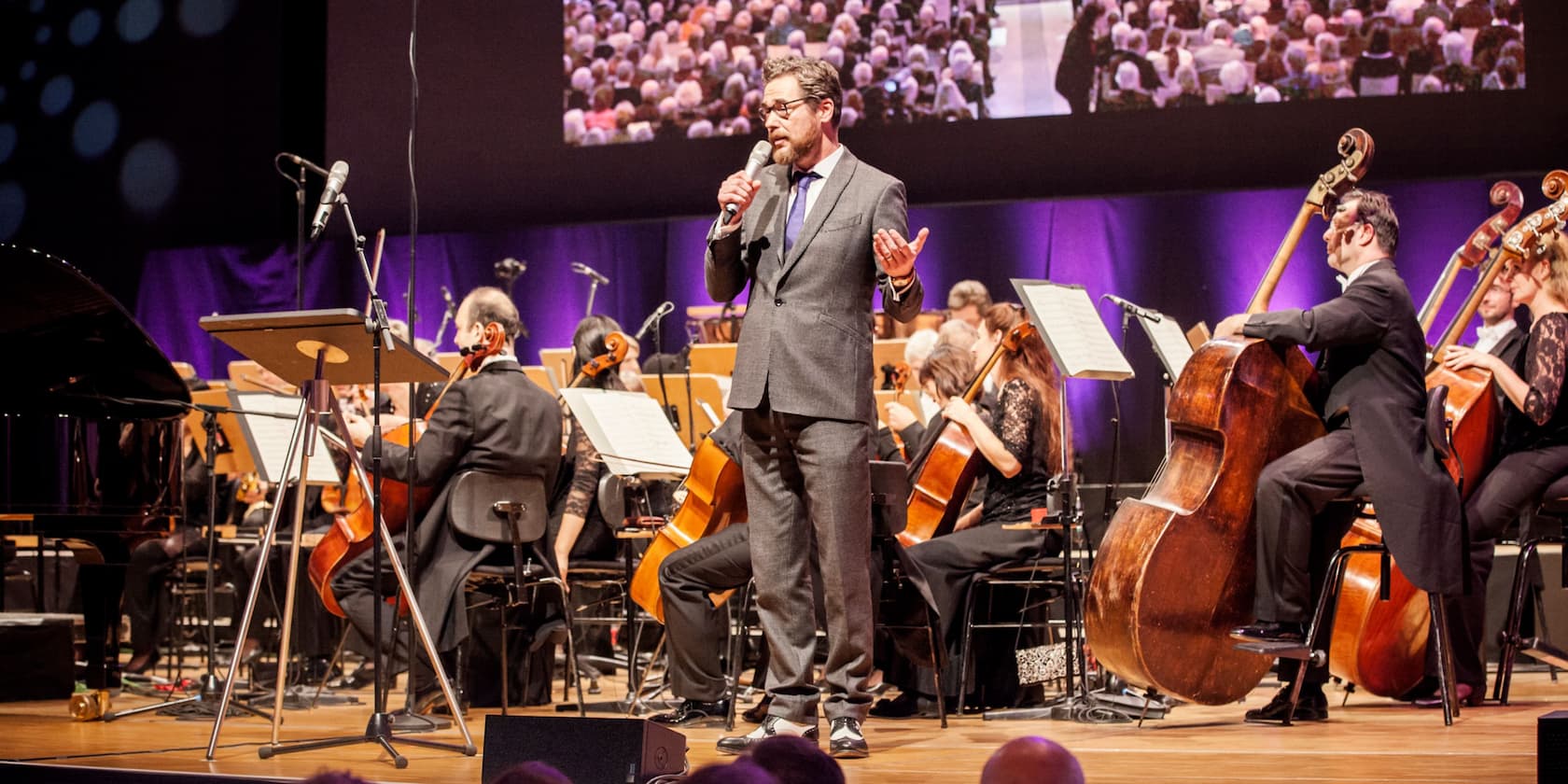 Ein Mann hält ein Mikrofon und spricht vor einem Orchester auf einer Bühne.