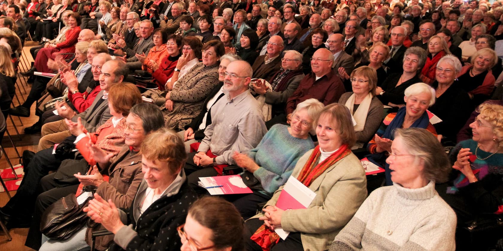 Eine große Gruppe von Menschen sitzt in einem Auditorium und applaudiert, die meisten wirken älter.