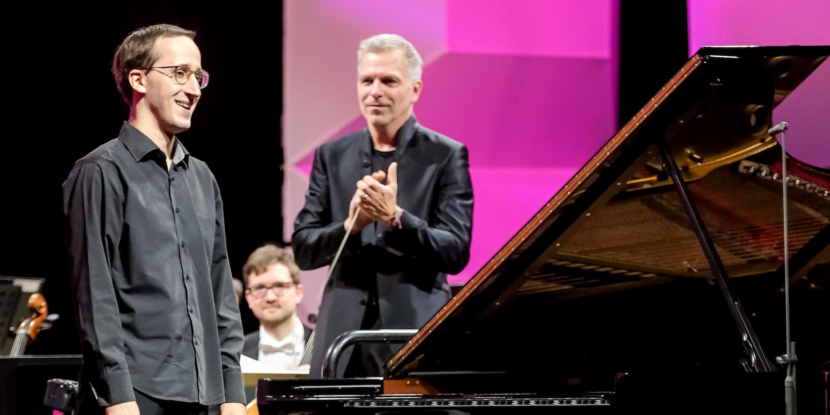 Ein Musiker steht neben einem Flügel auf einer Bühne, während ein Mann im Hintergrund applaudiert.