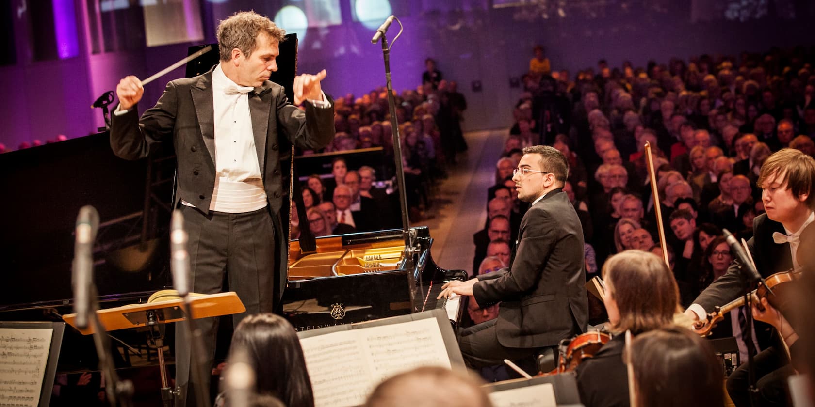 Dirigent und Pianist bei einem Konzert mit Orchester und Publikum im Hintergrund.
