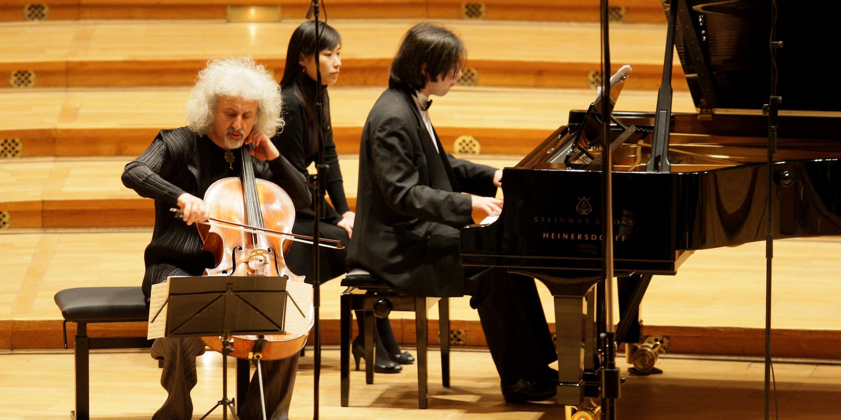 A cellist and a pianist performing on a concert stage, a woman observing in the background.
