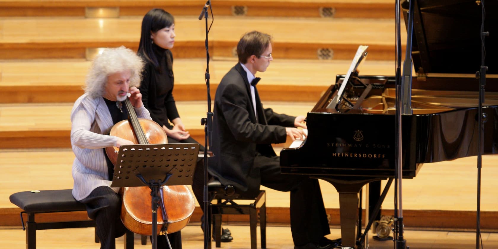 A cellist and a pianist performing on stage, with a woman sitting beside them.