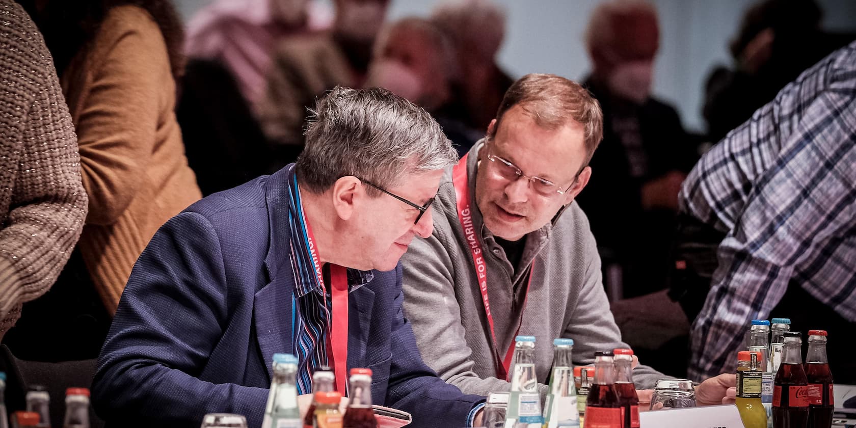 Zwei Männer unterhalten sich während einer Konferenz, umgeben von Getränkeflaschen auf einem Tisch.