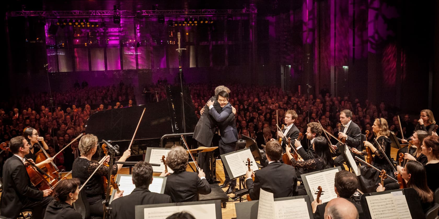 Dirigenten und Pianisten umarmen sich vor einem Orchester und einem applaudierenden Publikum.