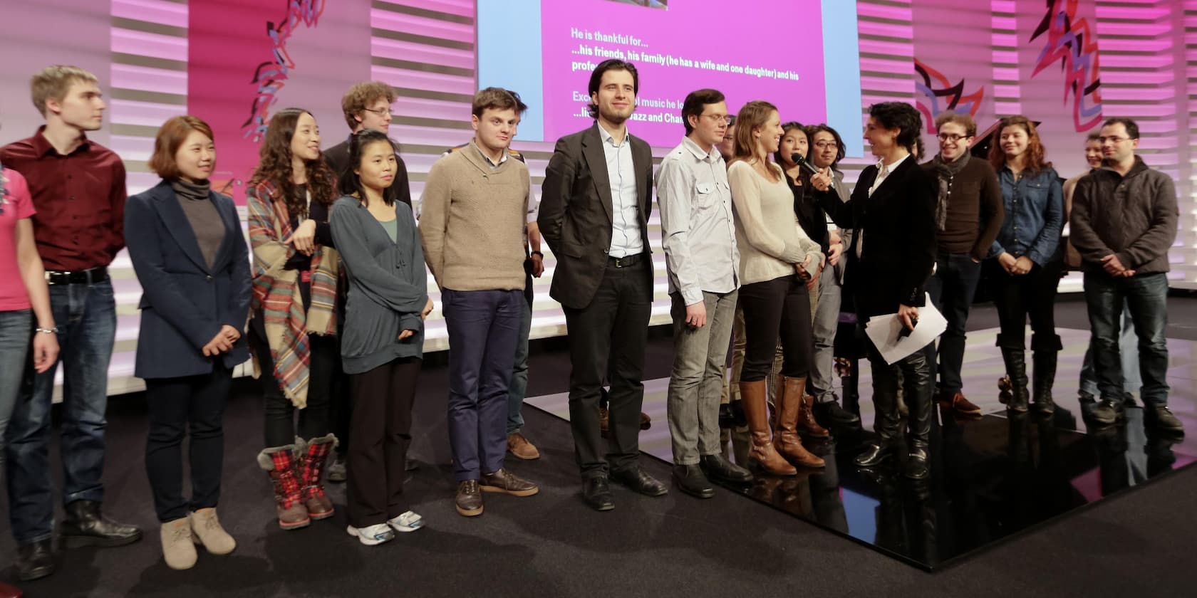 Eine Gruppe von Menschen steht auf einer Bühne vor einem rosa Hintergrund mit Text darauf.