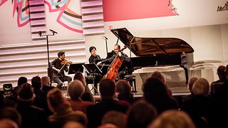 Im Fokus spielt ein Klaviertrio (Klavier, Violine, Violoncello) auf einer Bühne vor Publikum.