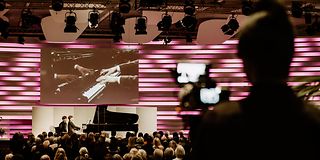 Blick in den Saal hinter dem sitzenden Publikum im Konzert. Ein Mann spielt Flügel, seine Hände sind auf dem Bildschirm über ihm