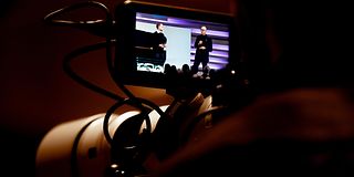 Kameraobjektiv im Dunkeln. Zwei Männer in dunkler Kleidung sprechen auf der Bühne vor magentafarbener Wand.