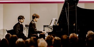 Zweigeteiltes Bild: Unten Pianist mit Notenwender am Flügel auf der Bühne, oben die beiden auf rosa Screen.
