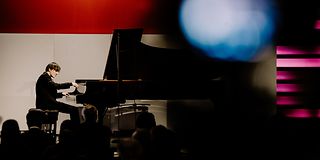Ein Pianist mit halblangem, dunkelblondem Haar spielt auf der Bühne am Flügel. Die rechte Bildseite ist verschwommen.