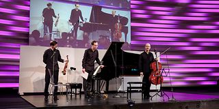 Ein Geiger, ein Pianist und ein Cellist, alle in schwarzer Kleidung, verbeugen sich auf der Bühne.