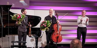 Der Geiger und Cellist stehen mit Blumensträußen lächelnd auf der Bühne. Der Moderator rechts neben ihnen applaudiert.