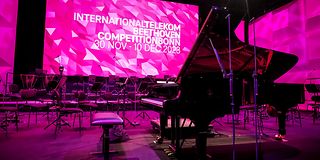 Auf der menschenleeren Bühne sind Orchester und Flügel aufgebaut, dahinter der magenta Screen mit Logo des Wettbewerbs.