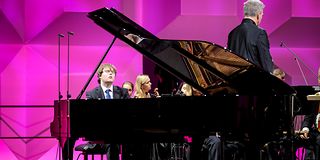 Ein Pianist im schwarzem Anzug und blauer Krawatte spielt emotional mit geschlossenen Augen. Rechts daneben der Dirigent.