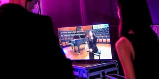 Im Backstage stehen zwei Menschen vor einem Bildschirm, der Zhouhui Shen im Kammermusiksaal zeigt.