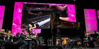 Eine Pianistin im Pailettenkleid spielt mit schwungvoller Geste am Flügel auf der Bühne mit dem Orchester.