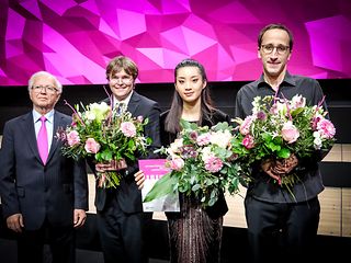 Rechts neben dem weißhaarigen Jurypräsident stehen die drei Preisträger:innen mit großen Blumensträußen vor einer magenta Wand.