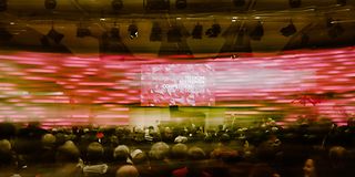 The audience sits in a hall and looks at a stage with a grand piano in front of a magenta glowing wall. Horizontally blurred.