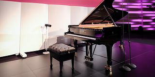 Opened grand piano with piano stool on the stage in front of a magenta illuminated wall and a banner with the competition logo.