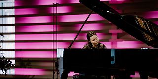 A woman with dark hair tied back plays on an open grand piano in front of a magenta-lit wall.