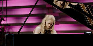 A woman with long blond hair plays emotionally on an open grand piano in front of a magenta-lit wall.