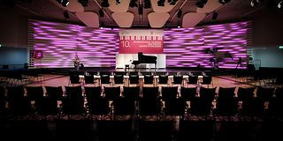 An empty, dark hall with rows of chairs, a long jury table and a stage with a black grand piano and magenta-lit wall.