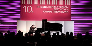 In the dark hall, a man with his black tied up hair plays in front of an audience on a grand piano in front of a magenta wall.