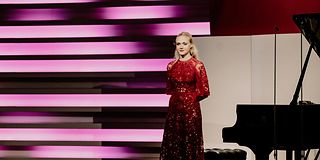 A blonde woman in a glittering red long dress stands in front of the grand piano with her arms crossed behind her back.
