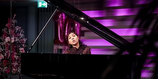 A young woman with curly black hair can be seen frontally through the open grand piano. 
