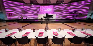 An empty long table with magenta folders on each seat stands in front of the empty stage.
