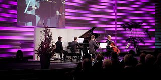 Three musicians in piano trio playing on stage with magenta background. The viewing angle is to the left of the stage.
