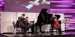 A piano trio plays on stage, and on the screen above you can see the pianist's hands.