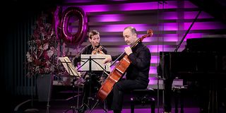 A violinist and a cellist play expressively while seated on the magenta-lit stage.