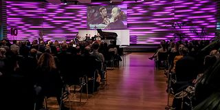 In the full hall, the view goes from behind the fully occupied audience to the magenta stage with the musicians on the screen.