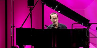 A young pianist in glasses and a black shirt plays with a concentrated face in front of magenta walls.