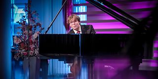 A young man with glasses and brown hair plays emotionally on the grand piano. Under the wing, the colors blur. 