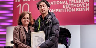 A middle-aged woman poses with a black-haired pianist and the certificate for a prize won.