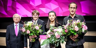 To the right of the jury president, the three award winners stand in front of a magenta wall with large bouquets of flowers.