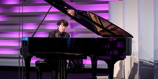A young black-haired man plays emotionally on the grand piano with his eyes closed.