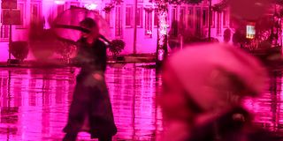 Through a magenta shimmering glass wall, you can see a woman walking outside with an umbrella from the inside.