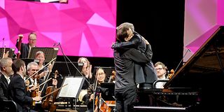 A young pianist and the conductor embrace each other on stage in front of the seated orchestra.