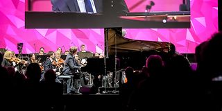 A young pianist plays on stage with orchestra in front of magenta-lit walls.