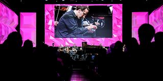 A young pianist plays on stage with orchestra in front of magenta-lit walls in the dark, packed hall.