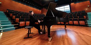 A black-haired woman in black clothes plays with her back straight at the grand piano in an empty hall.
