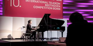 A man with black hair plays on the grand piano on stage in front of a magenta banner with white lettering.