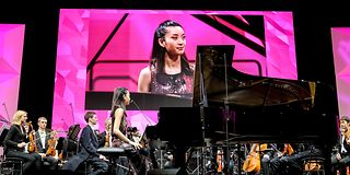 A pianist in a sequined dress sits at the grand piano and looks at the conductor. Her face can be seen on the screen.
