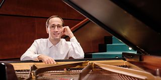 A brown-haired man with glasses in a white shirt rests his head on one hand and smiles over the taut strings of the grand piano